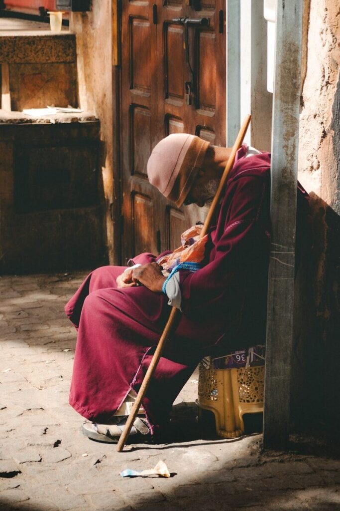 Elderly man in maroon robe rests with cane.
